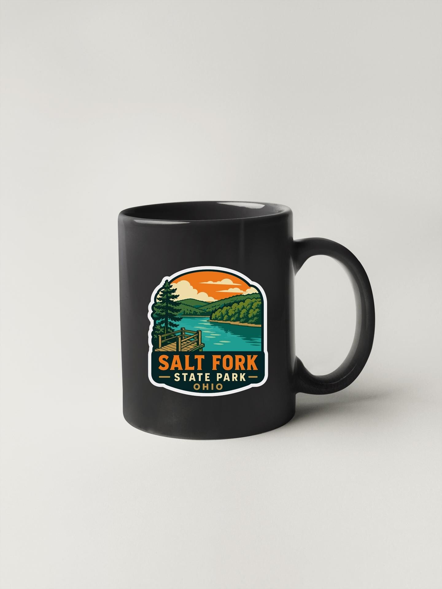 A black coffee mug with a graphic design depicting a scenic landscape, including a lake, mountains, and a sunset.