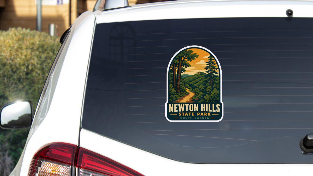A sticker on the rear window of a white car, depicting a scenic landscape with a winding road and trees, advertising Newton Hills State Park in South Dakota.