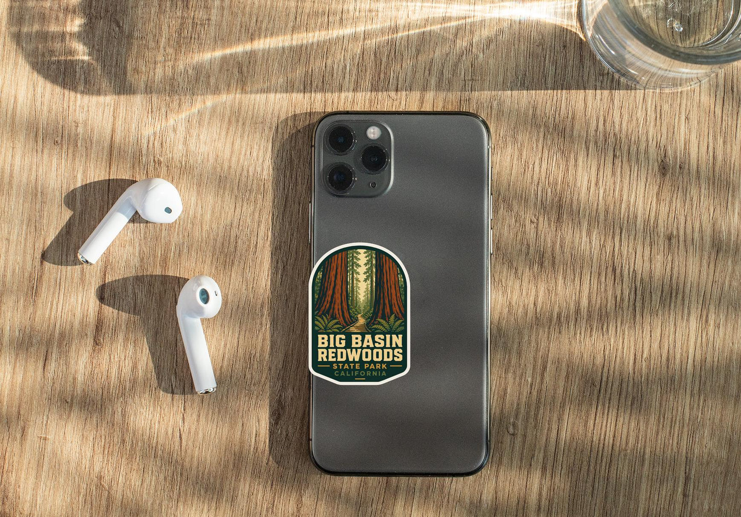 a smartphone with a sticker on it, placed on a wooden surface alongside a pair of white earbuds and a glass of water.