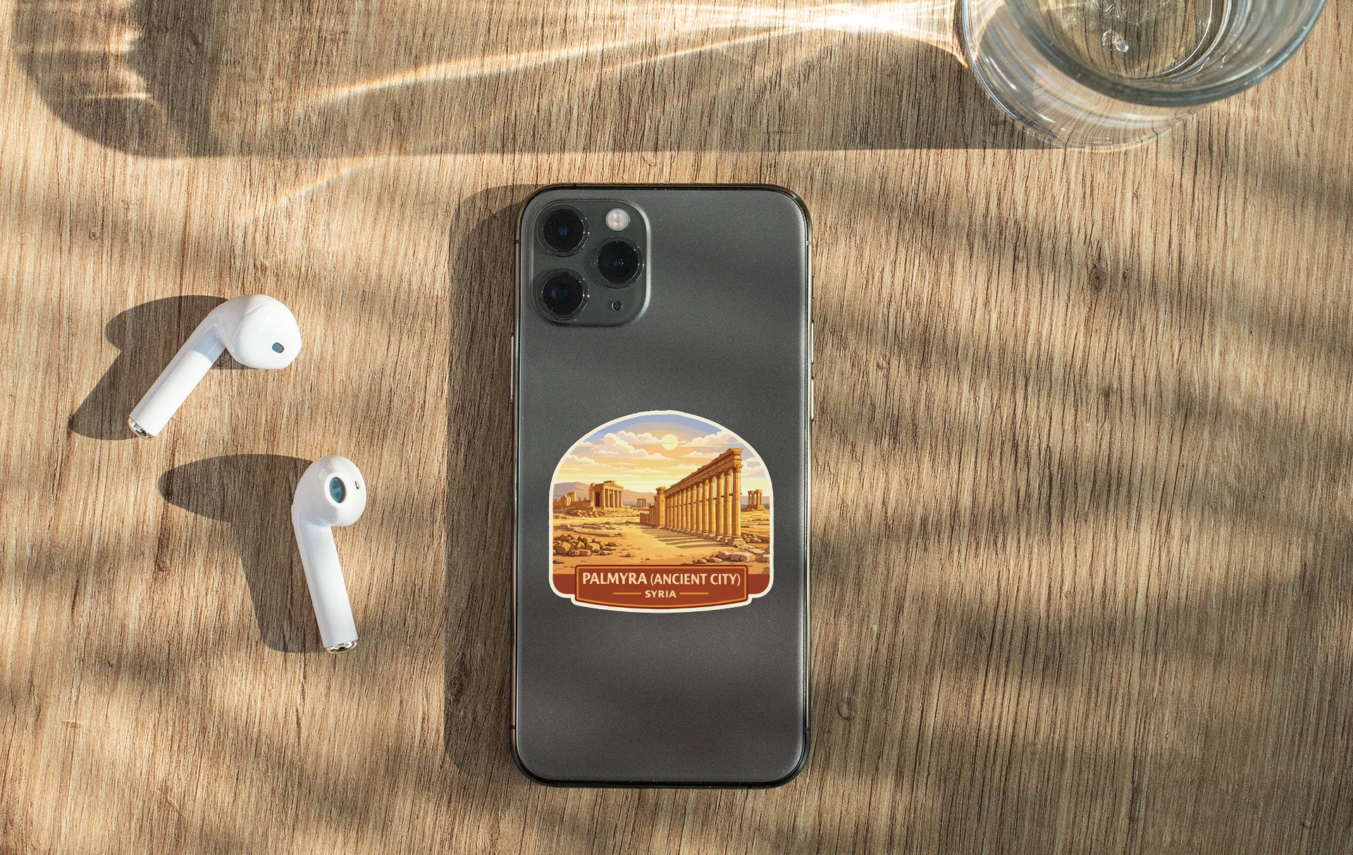 a smartphone with a sticker of a city on it, placed on a wooden surface alongside a pair of white earbuds and a glass of water.
