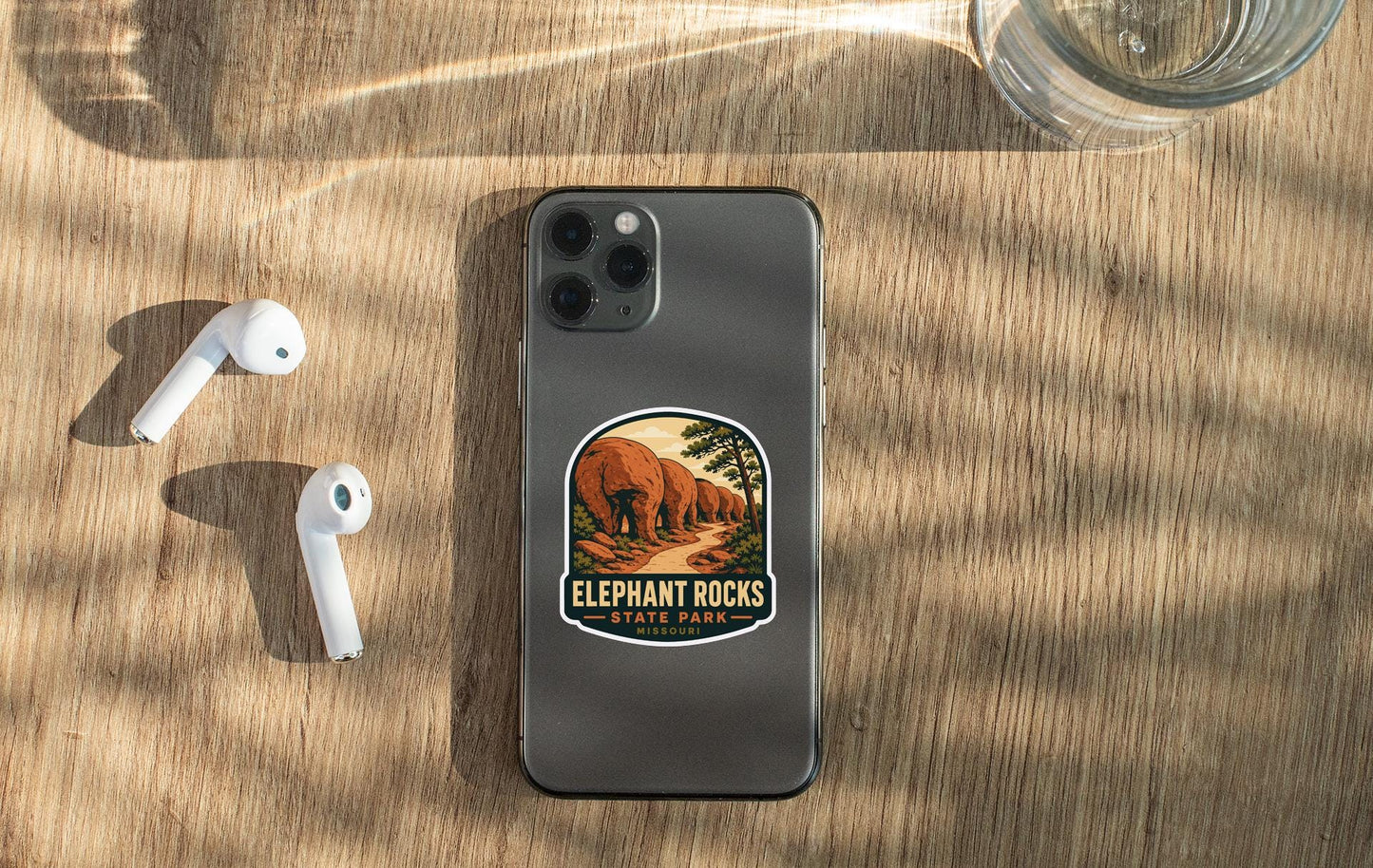 a smartphone with a sticker of Elephant Rocks State Park on its back, along with a pair of white earbuds and a glass of water on a wooden surface.