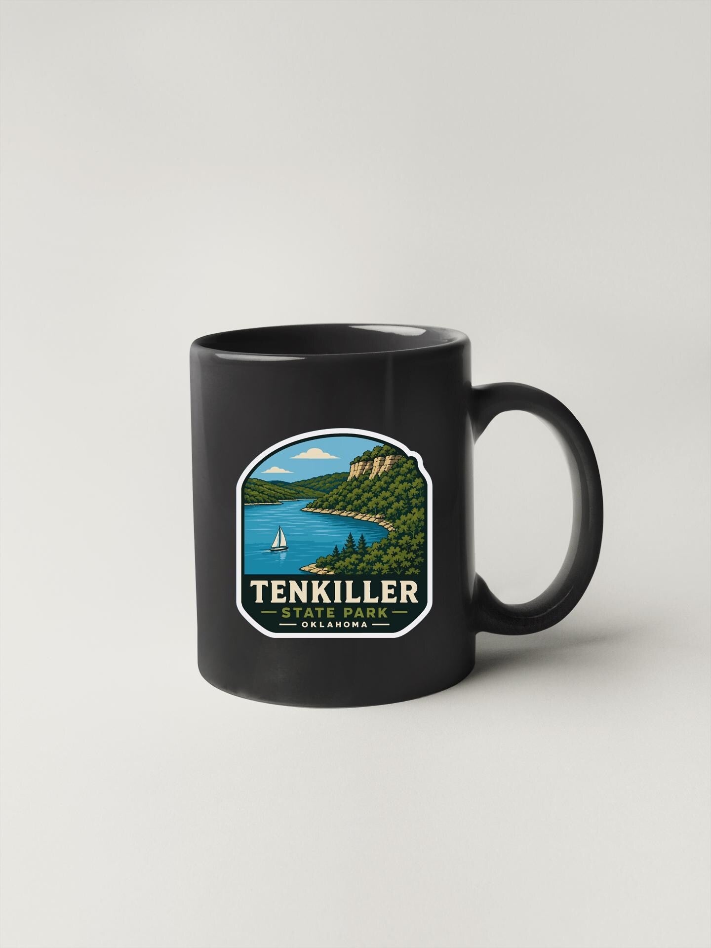 A black coffee mug with a graphic design depicting a scenic landscape, including a lake, mountains, and a sailboat.