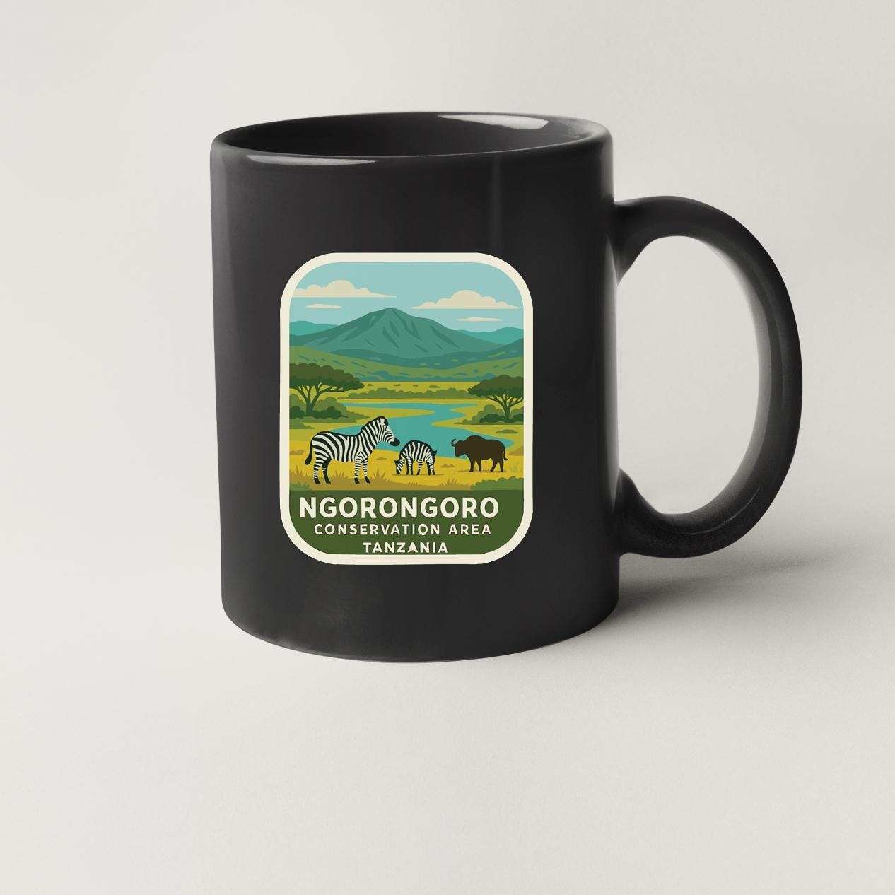 A black coffee mug with a label depicting a scenic landscape with zebras, a cow, and mountains in the background, along with the text "NGORONGOORO CONSERVATION AREA TANZANIA".