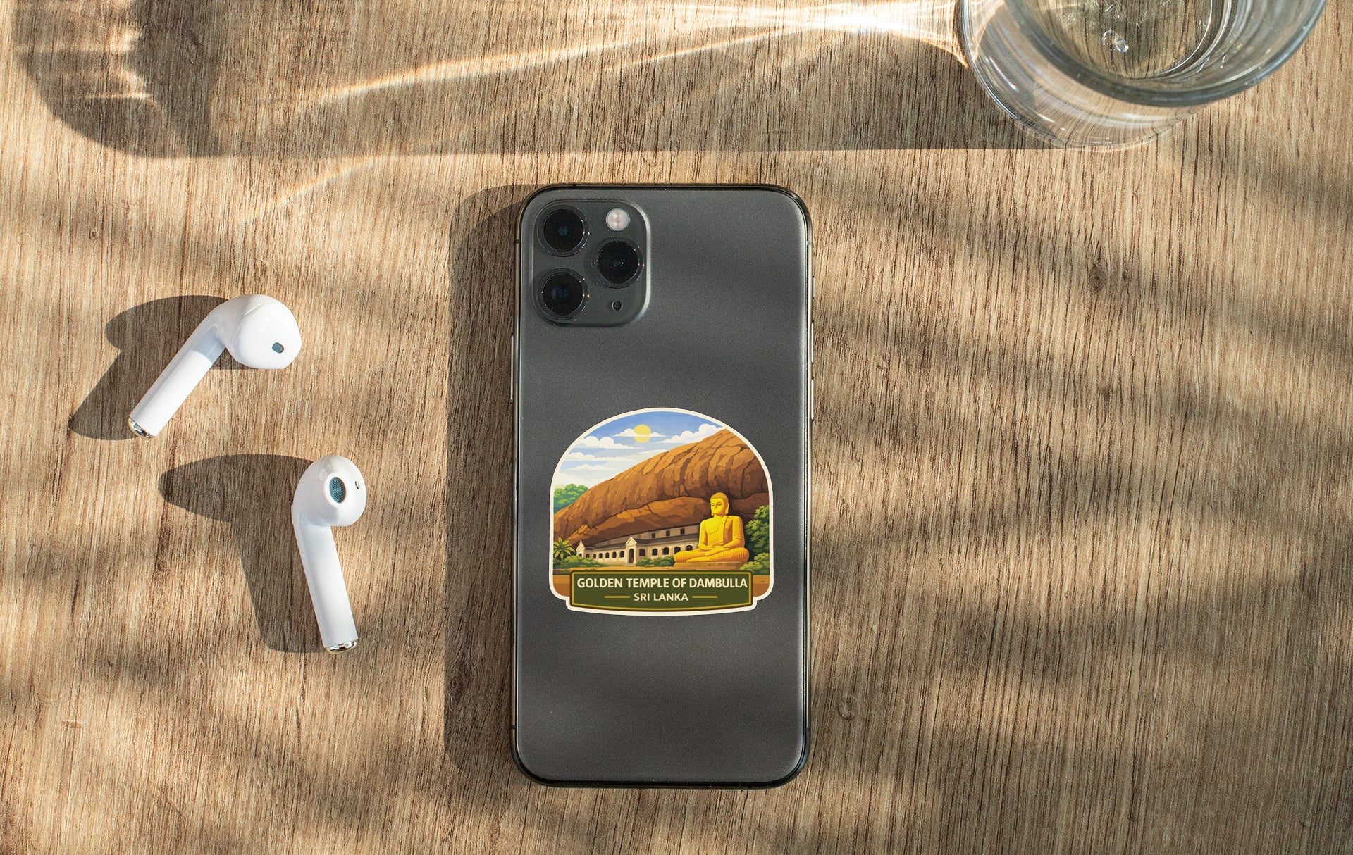 a smartphone with a sticker of the Golden Temple of Sambora, a famous Buddhist temple in Thailand, placed on a wooden surface alongside a pair of white earbuds and a glass of water.
