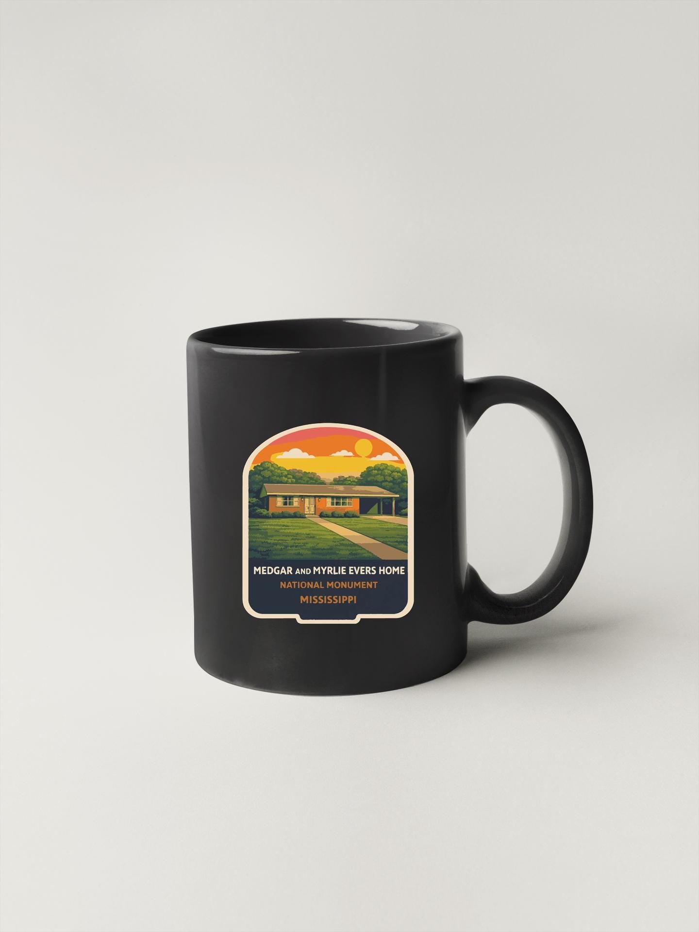 A black coffee mug with a graphic design depicting a house and trees, along with text that reads "MEDGAR AND MYRUE EYERS HOME NATIONAL MONUMENT PINELLIPINI MS".