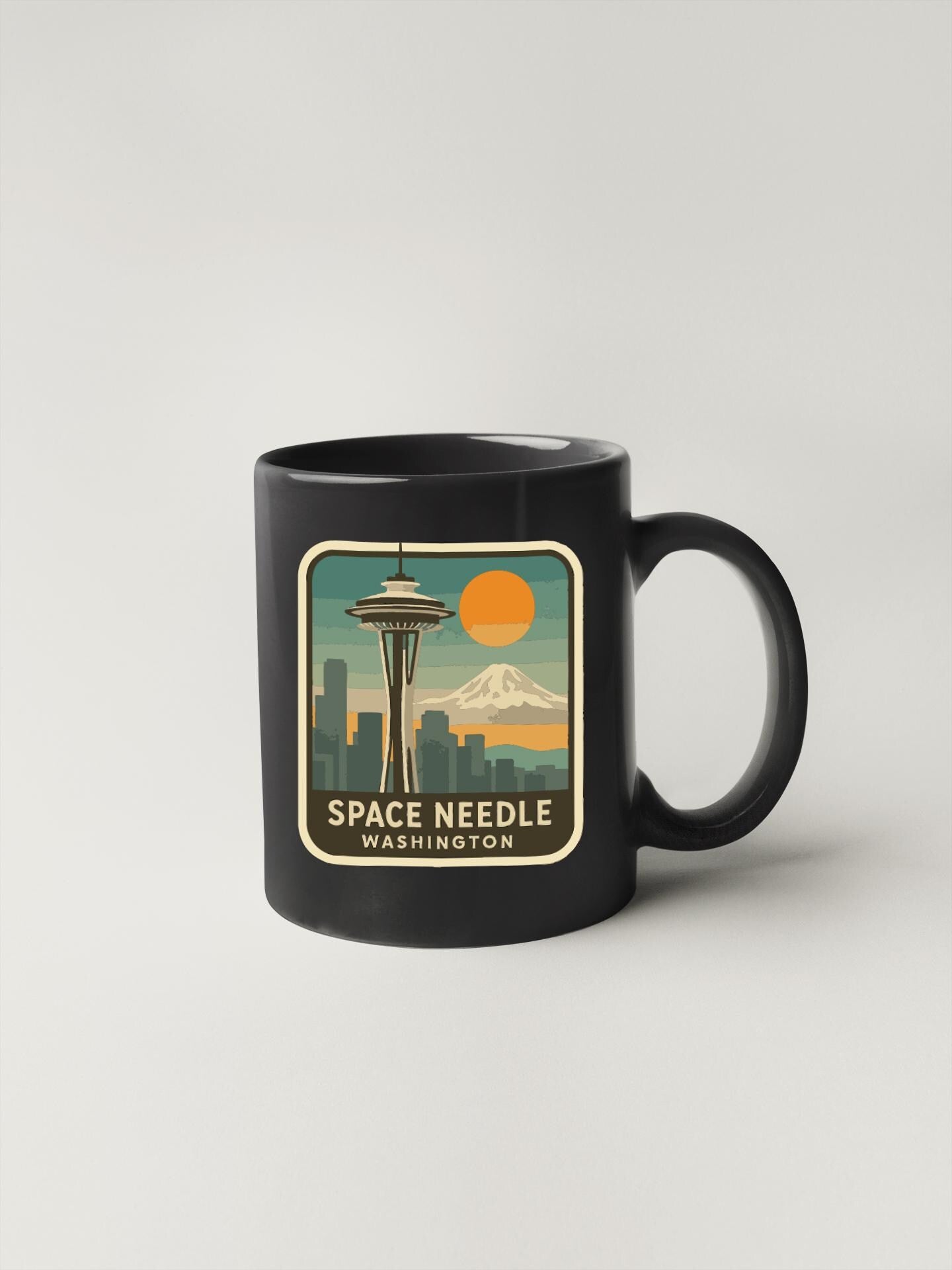 A black coffee mug with a graphic design featuring the Space Needle in Seattle, Washington, along with a mountain and a sunset.