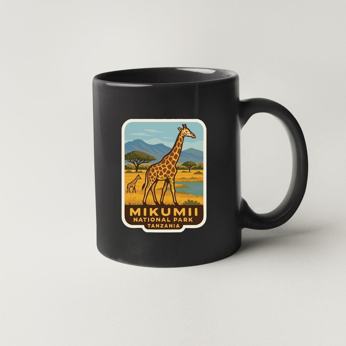 A black coffee mug with a graphic of a giraffe and the text "MIKUMII NATIONAL PARK TANZONIA" printed on it.