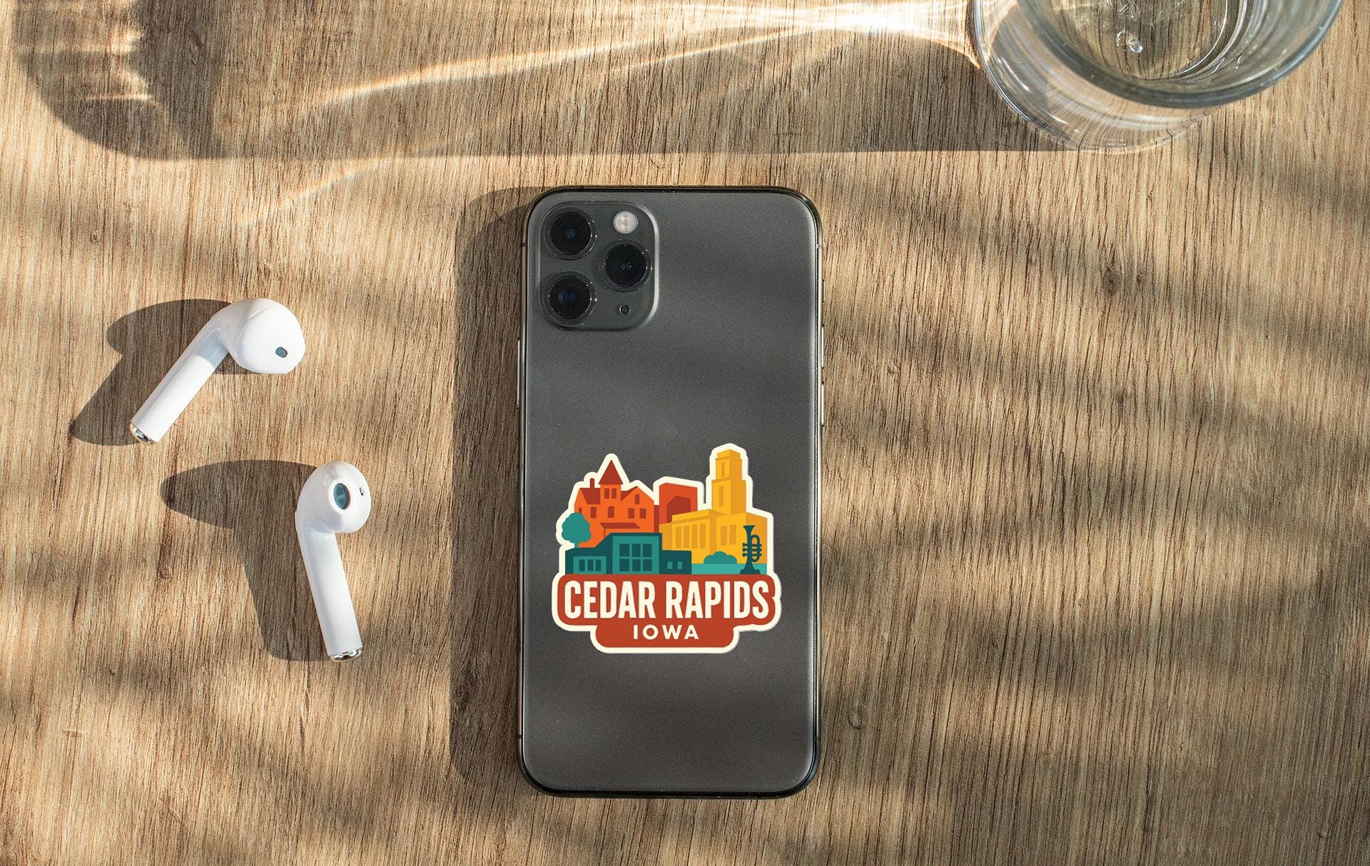 a smartphone with a sticker on the back, placed on a wooden surface alongside a pair of white earbuds and a glass of water.