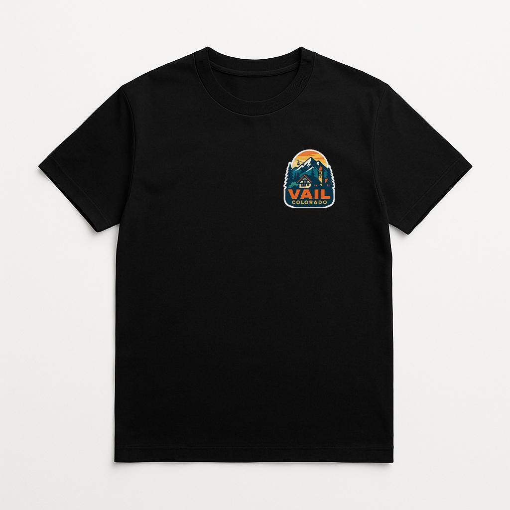 A black t-shirt with a graphic design on the front, featuring a mountain landscape with a house and the text "Vail" prominently displayed.