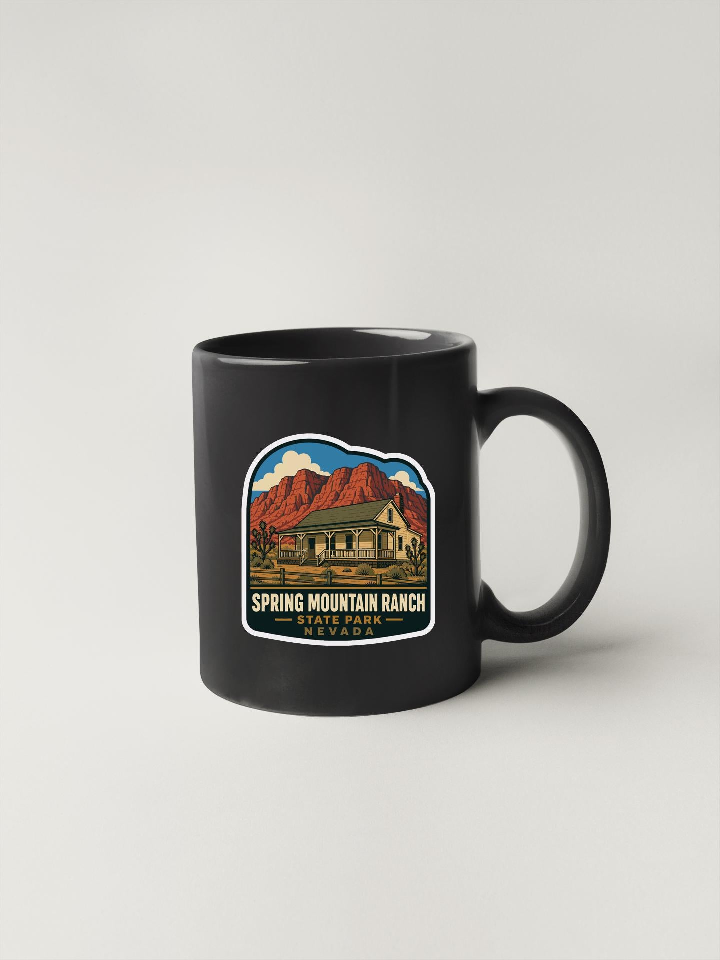 A black coffee mug with a graphic design depicting a house and mountains, along with the text "Spring Mountain Ranch State Park" printed on it.