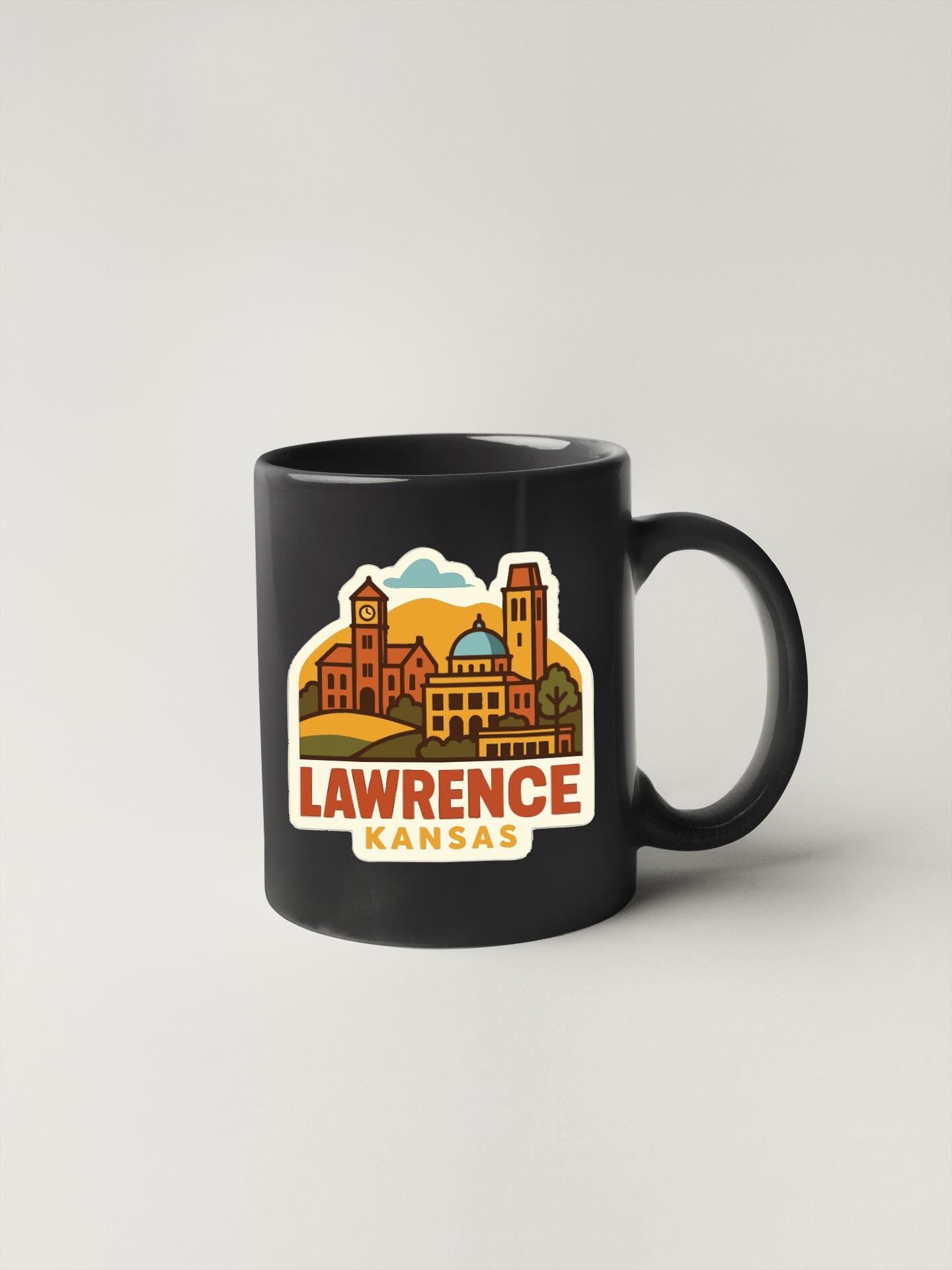 A black coffee mug with a colorful illustration of Lawrence, Kansas, featuring buildings, a clock tower, and a sunset.