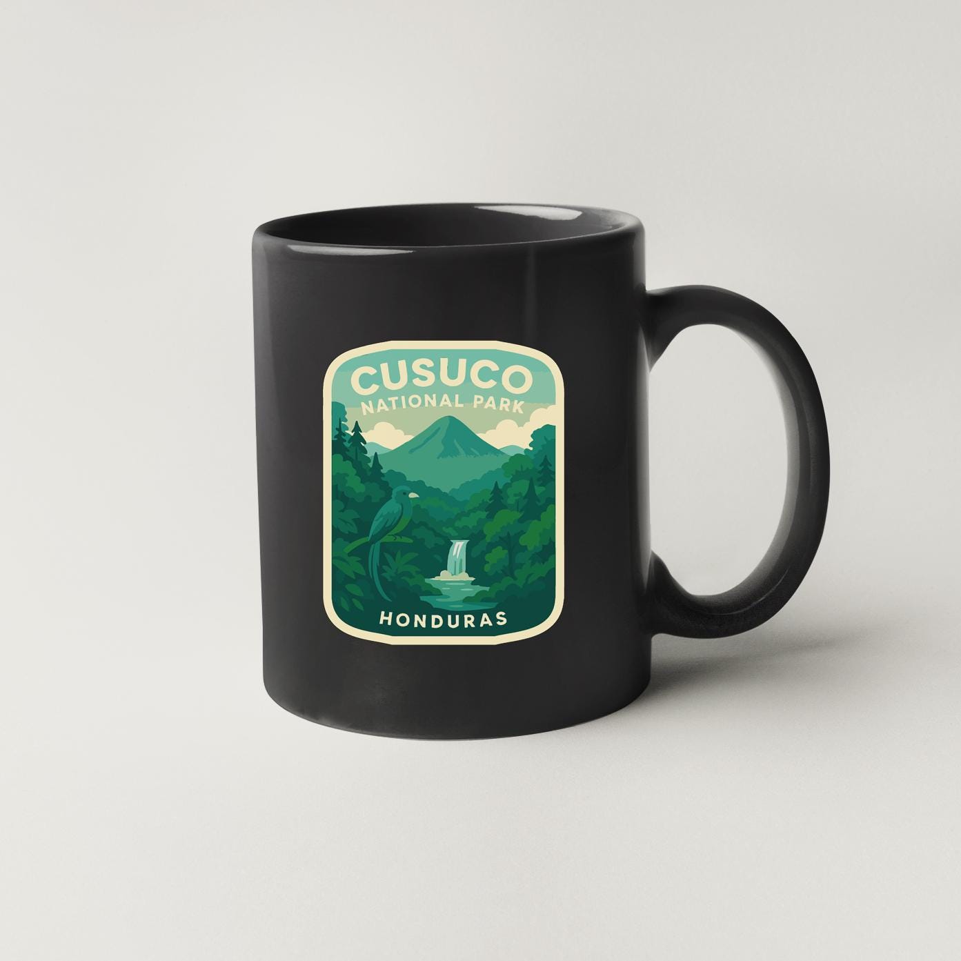 A black coffee mug with a graphic design depicting a tropical landscape, including a waterfall and a bird, along with the text "CUSUCO NATIONAL PARK HONDURAS".