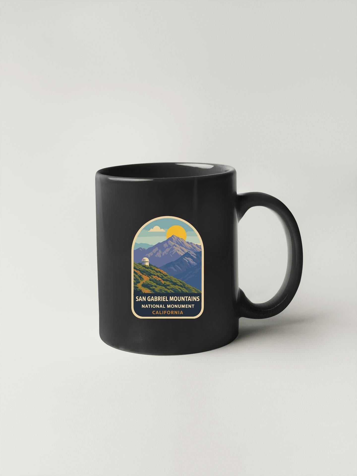 A black coffee mug with a graphic design depicting a mountain landscape and the text "San Gabriel Mountains National Monument California" printed on it.