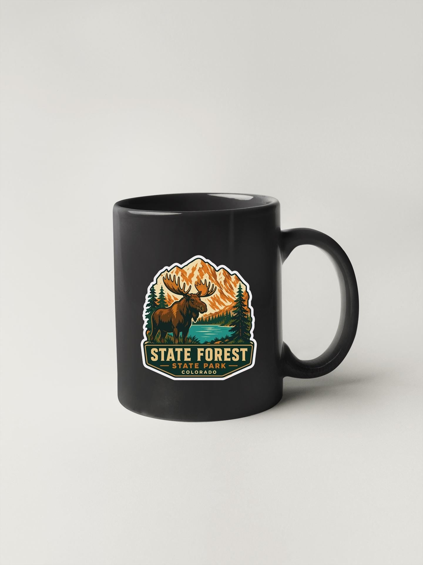 A black coffee mug with a graphic design depicting a moose and mountains, along with the text "STATE FOREST STATE PARK COLORADO".