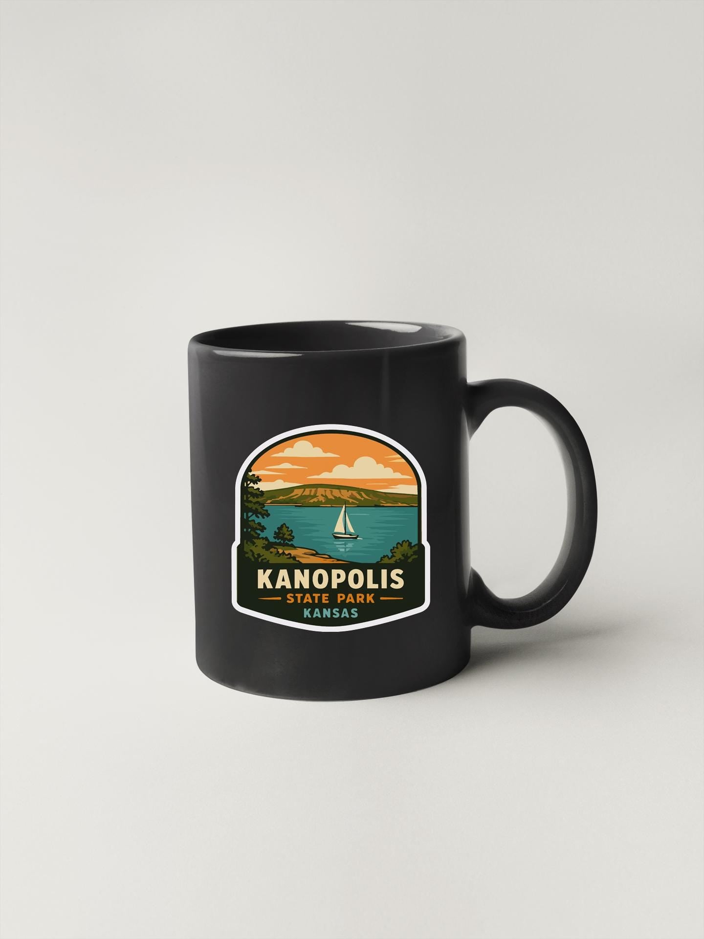 A black coffee mug with a sticker depicting a scenic landscape with a sailboat on the water.
