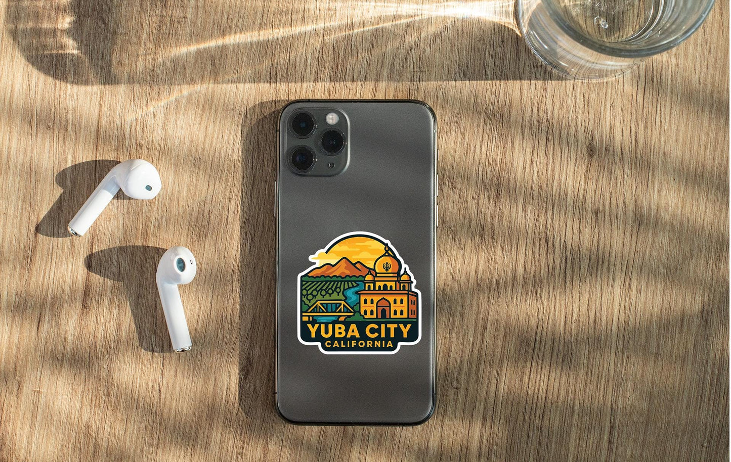 a smartphone with a sticker on it, placed on a wooden surface alongside a pair of white earbuds and a glass of water.
