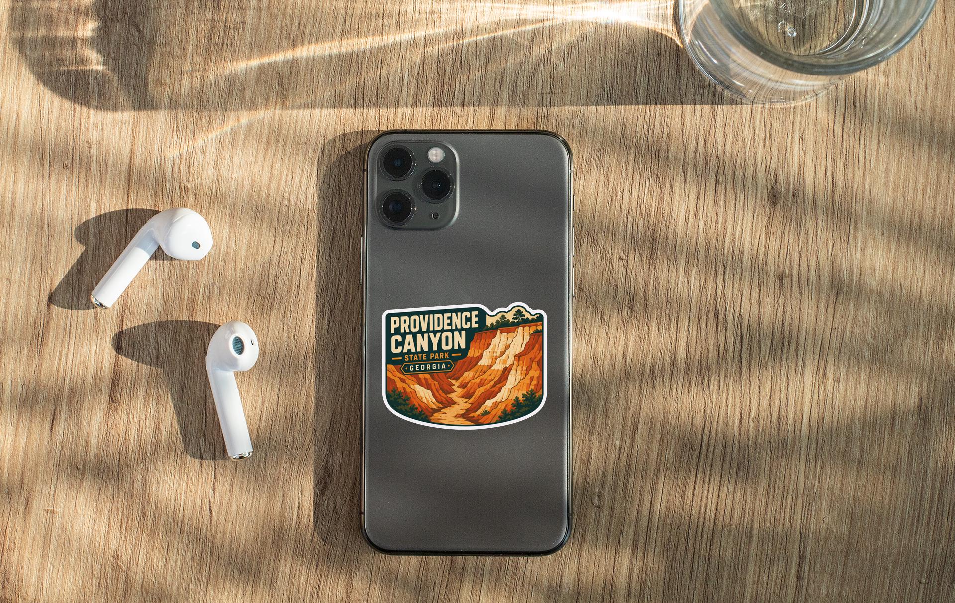 a smartphone with a sticker of Providence Canyon on it, along with a pair of white earbuds and a glass of water on a wooden surface.