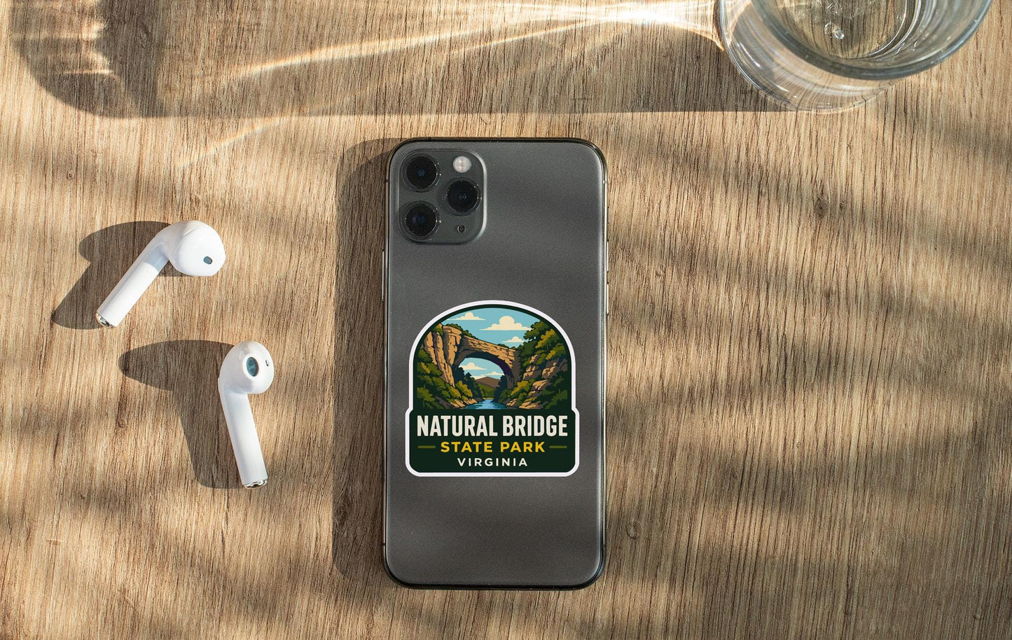 a smartphone with a sticker of Natural Bridge State Park Virginia on its back cover, along with a pair of white earbuds and a glass of water on a wooden surface.