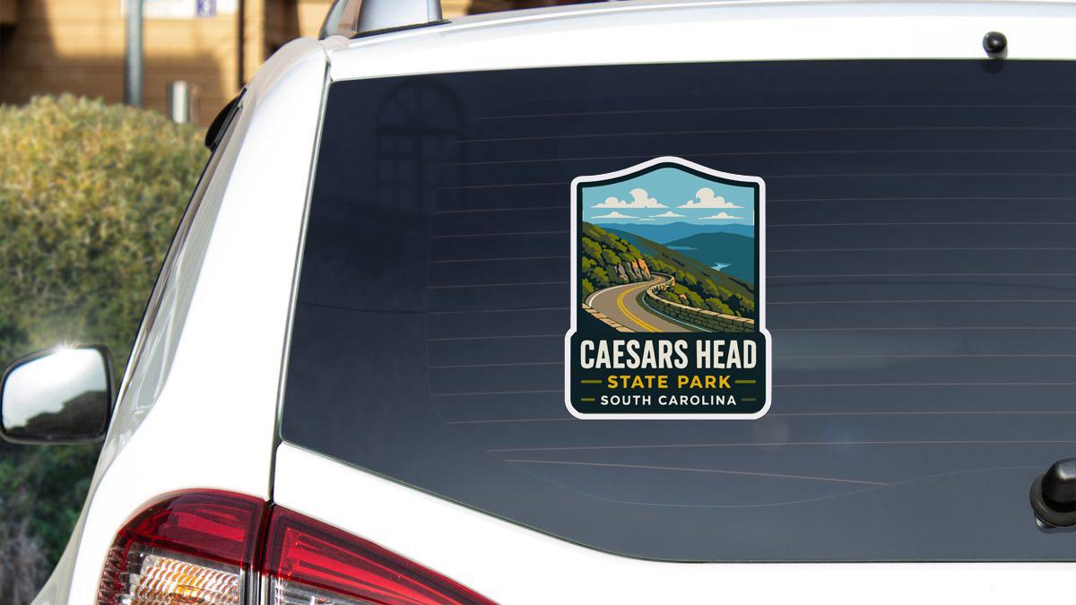 A sticker on the rear window of a white car, depicting a scenic landscape with a winding road and mountains in the background.