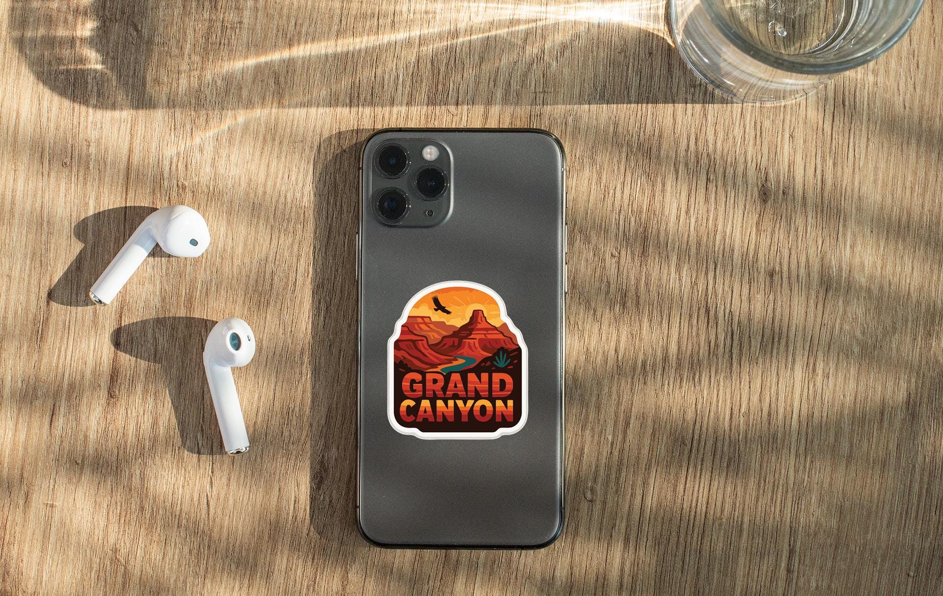 a smartphone with a sticker of the Grand Canyon on it, along with a pair of white earbuds and a glass of water on a wooden surface.