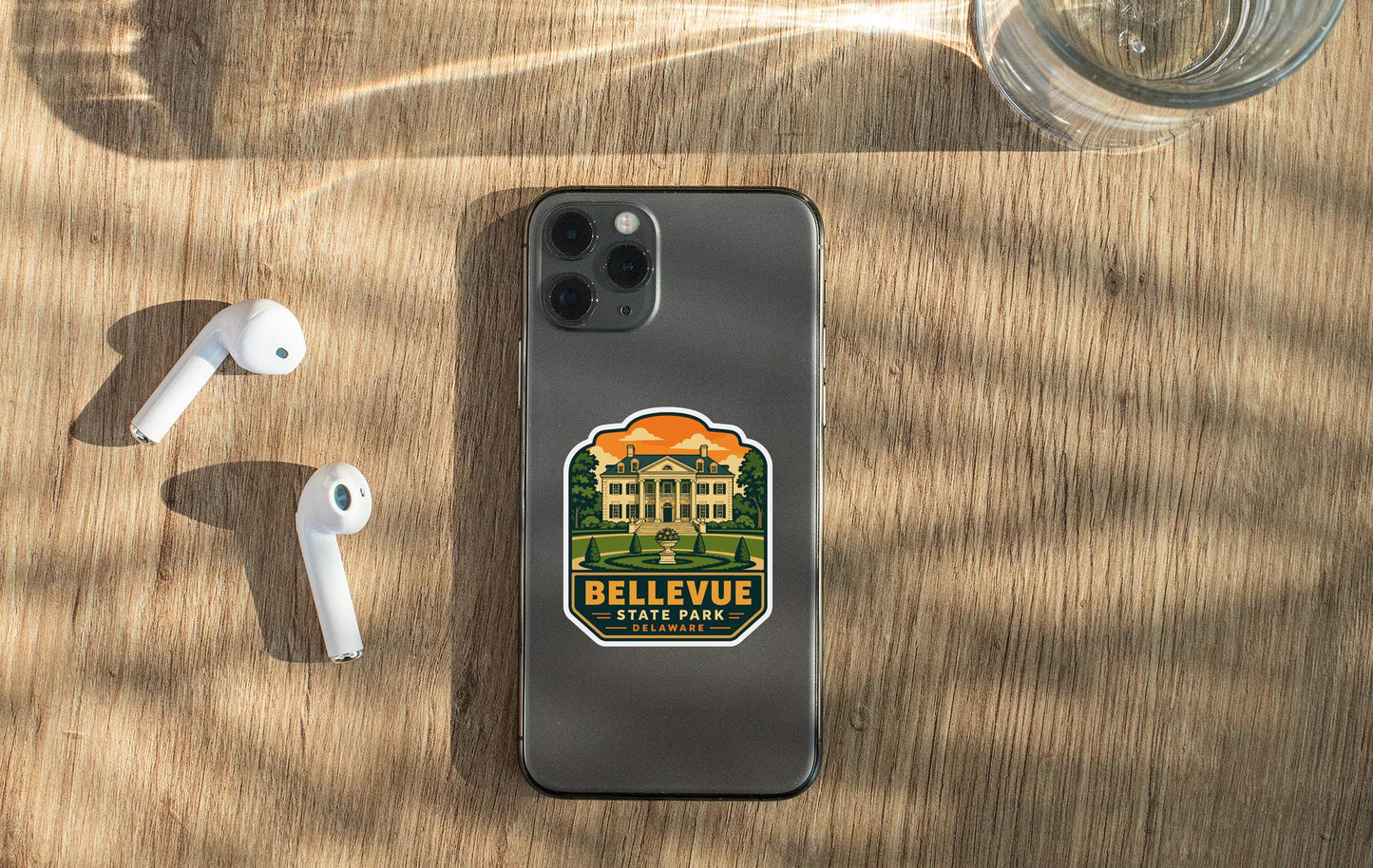 a smartphone with a sticker on the back, placed on a wooden surface alongside a pair of white earbuds and a glass of water.