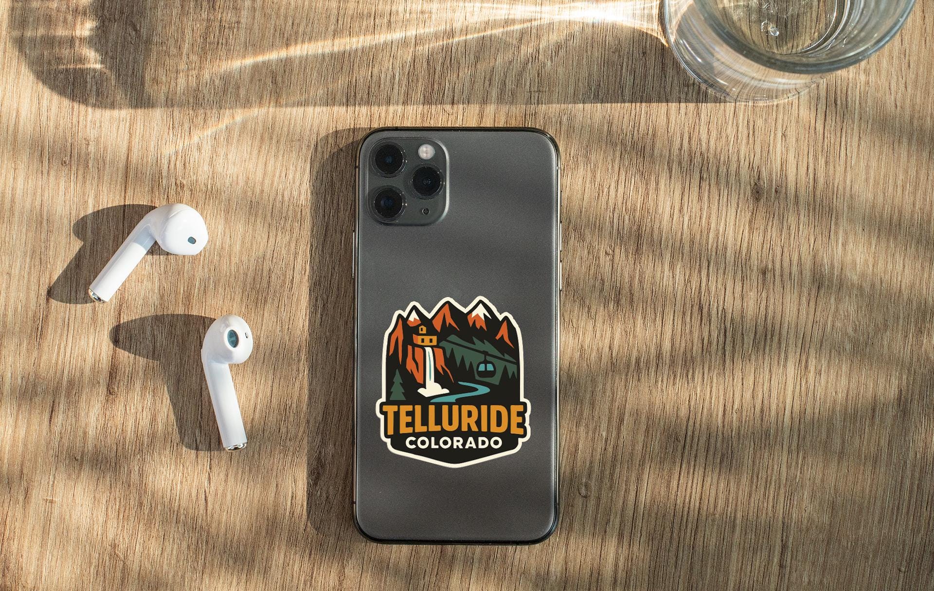 a smartphone with a sticker on the back, placed on a wooden surface alongside a pair of white earbuds and a glass of water.