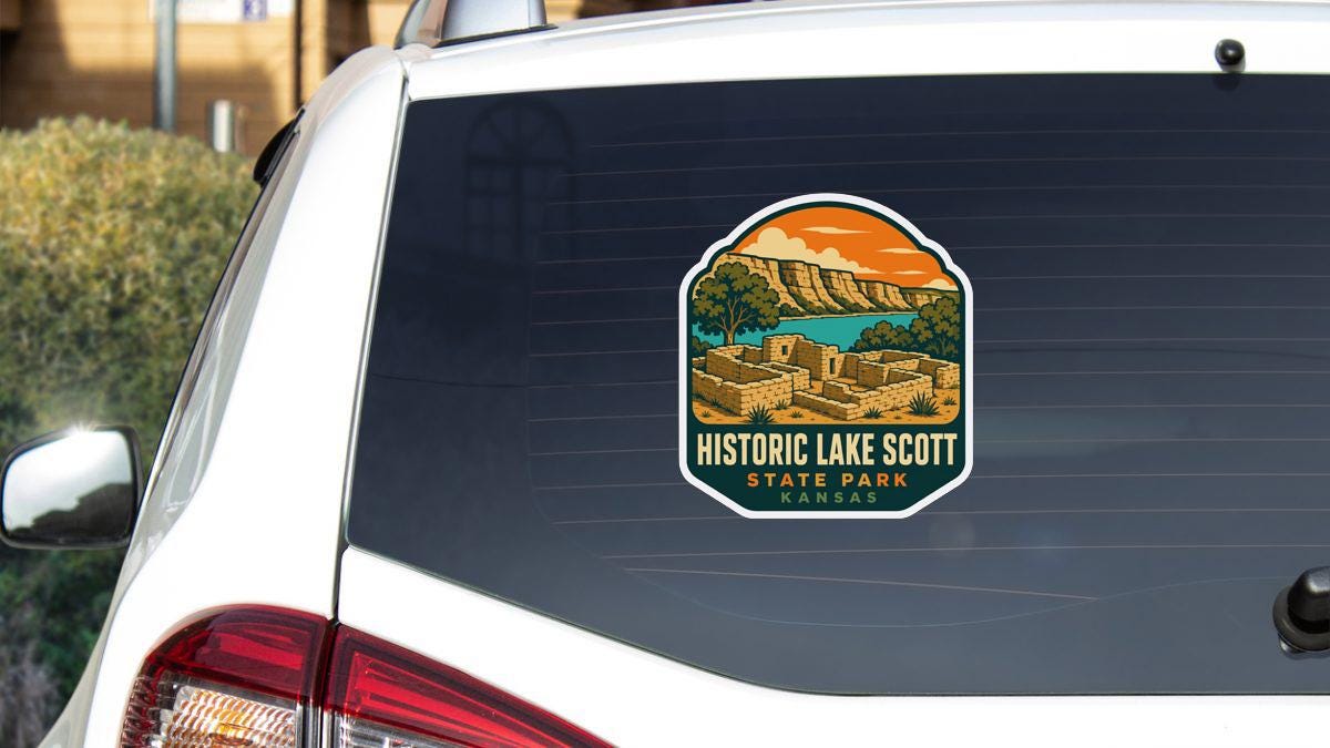 A sticker on the rear window of a white vehicle, depicting a scenic landscape with mountains and a lake, with the text "HISTORIC LAKE SCOTT STATE PARK KANSAS" printed on it.