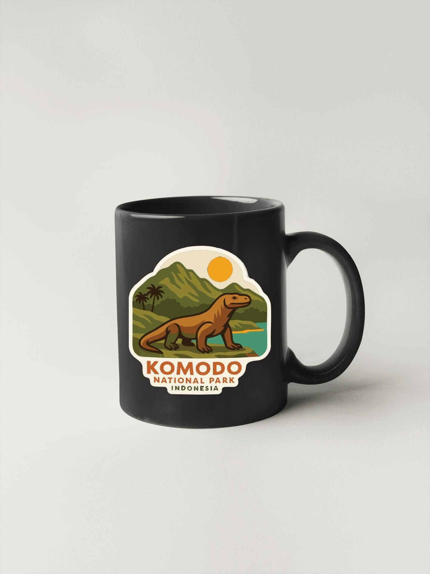 A black coffee mug with a sticker depicting a Komodo dragon in a tropical landscape.