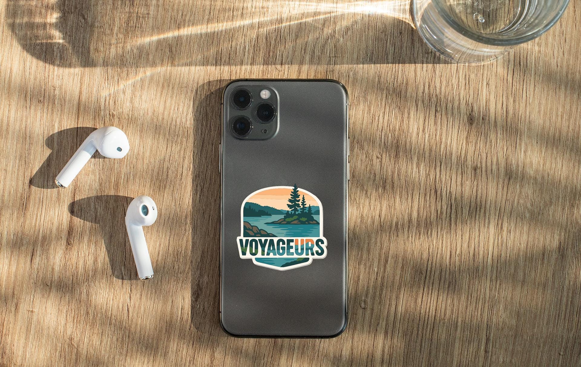 a smartphone with a sticker on the back, along with a pair of white earbuds and a glass of water on a wooden surface.