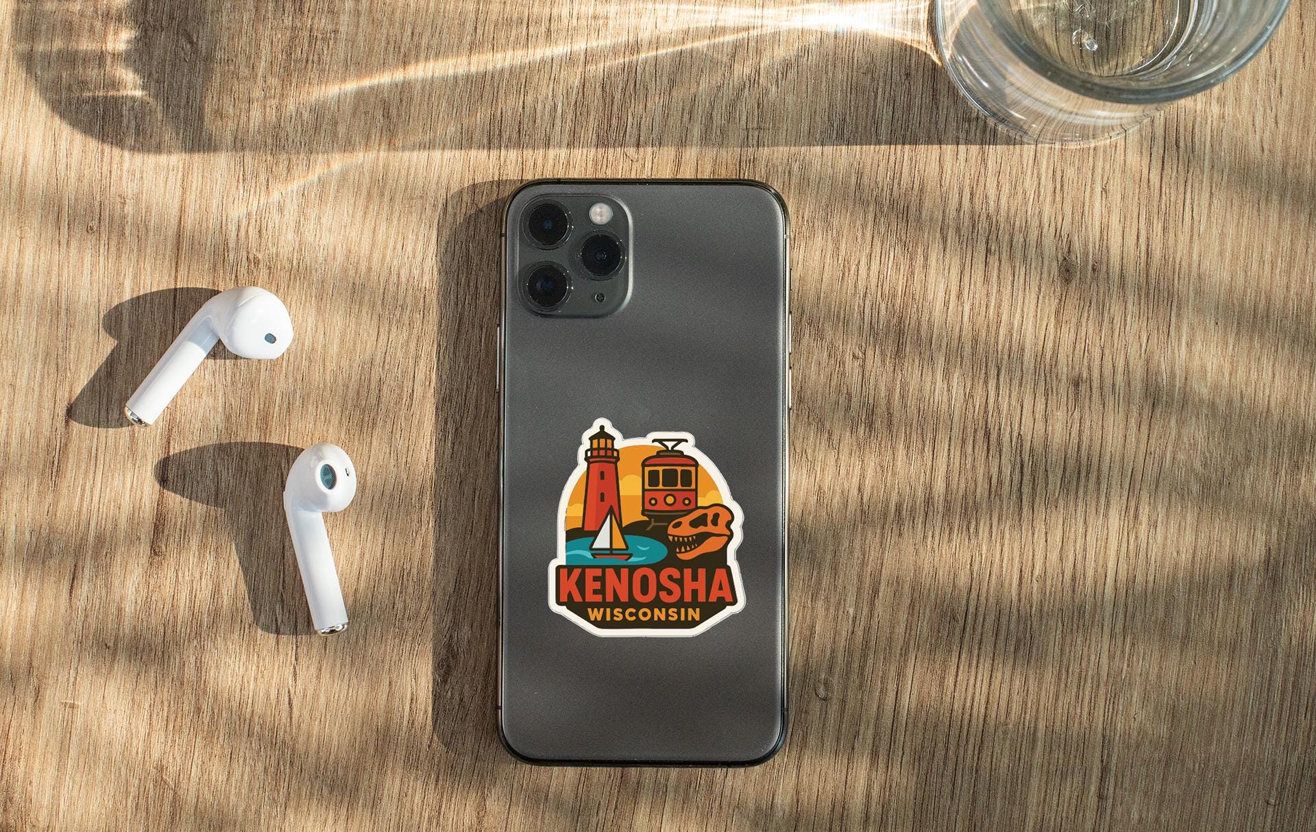 a smartphone with a sticker on the back, placed on a wooden surface alongside a pair of white earbuds and a glass of water.