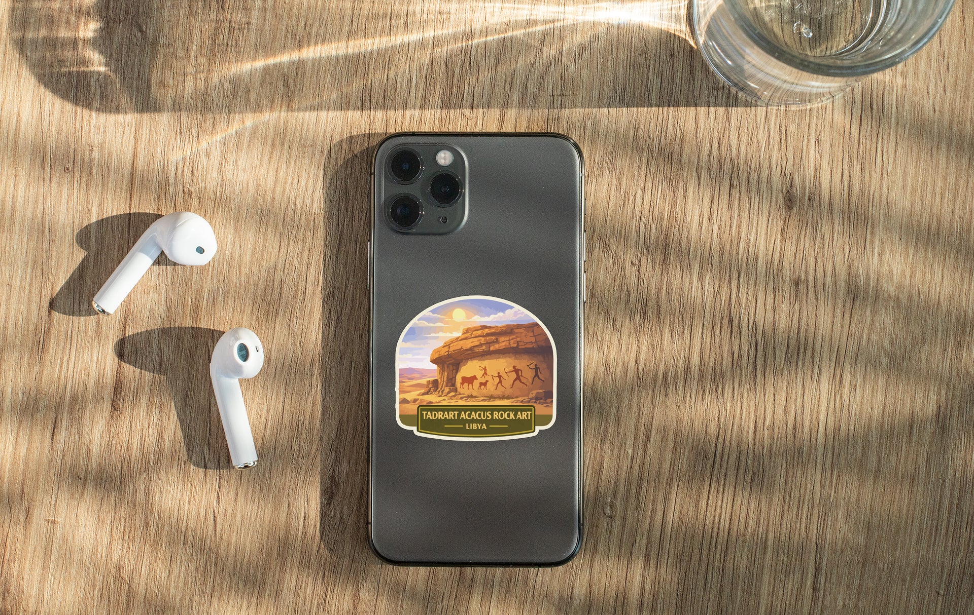 a smartphone with a sticker on the back, along with a pair of white earbuds and a glass of water on a wooden surface.