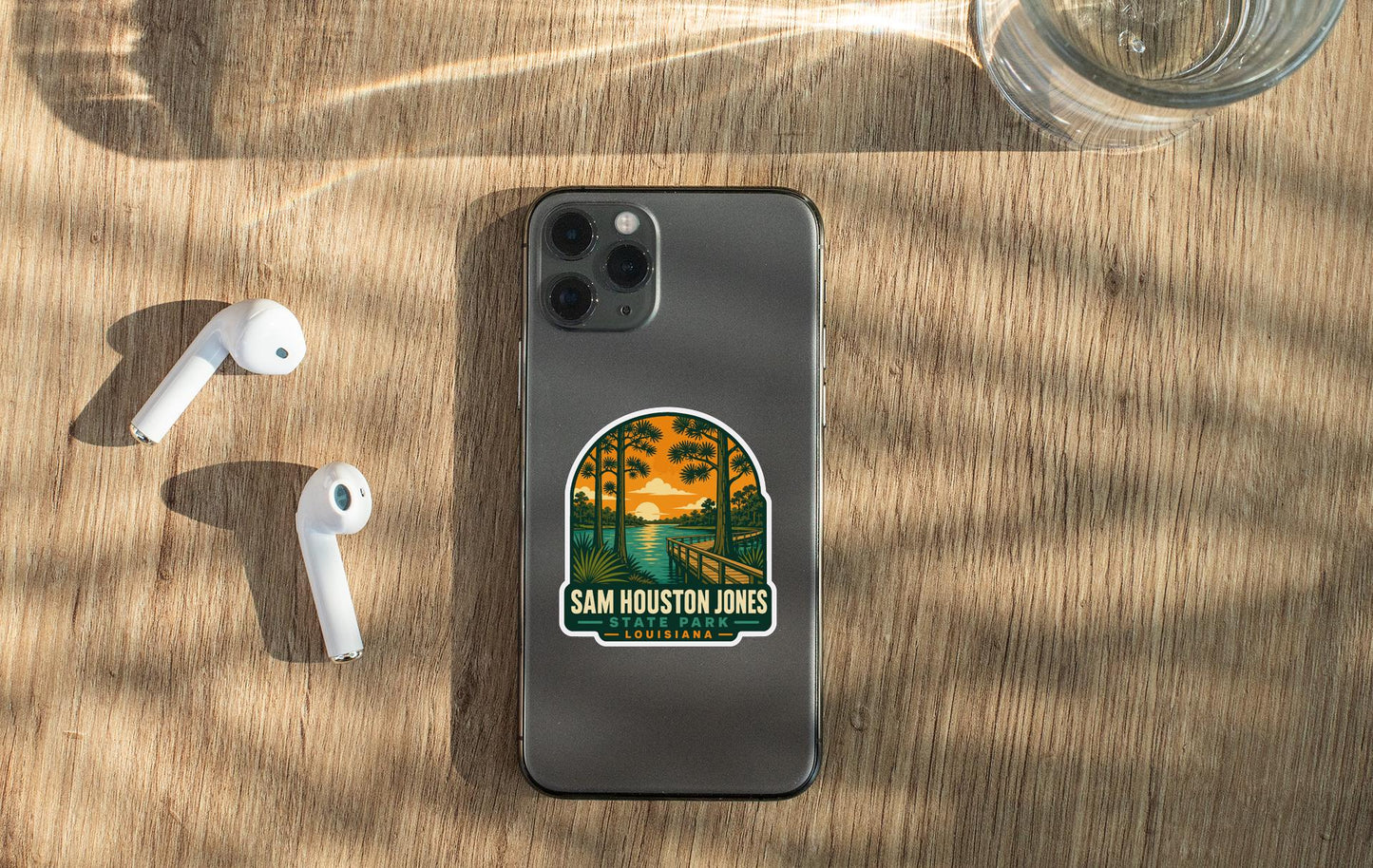 a smartphone with a sticker on the back, placed on a wooden surface alongside a pair of white earbuds and a glass of water.