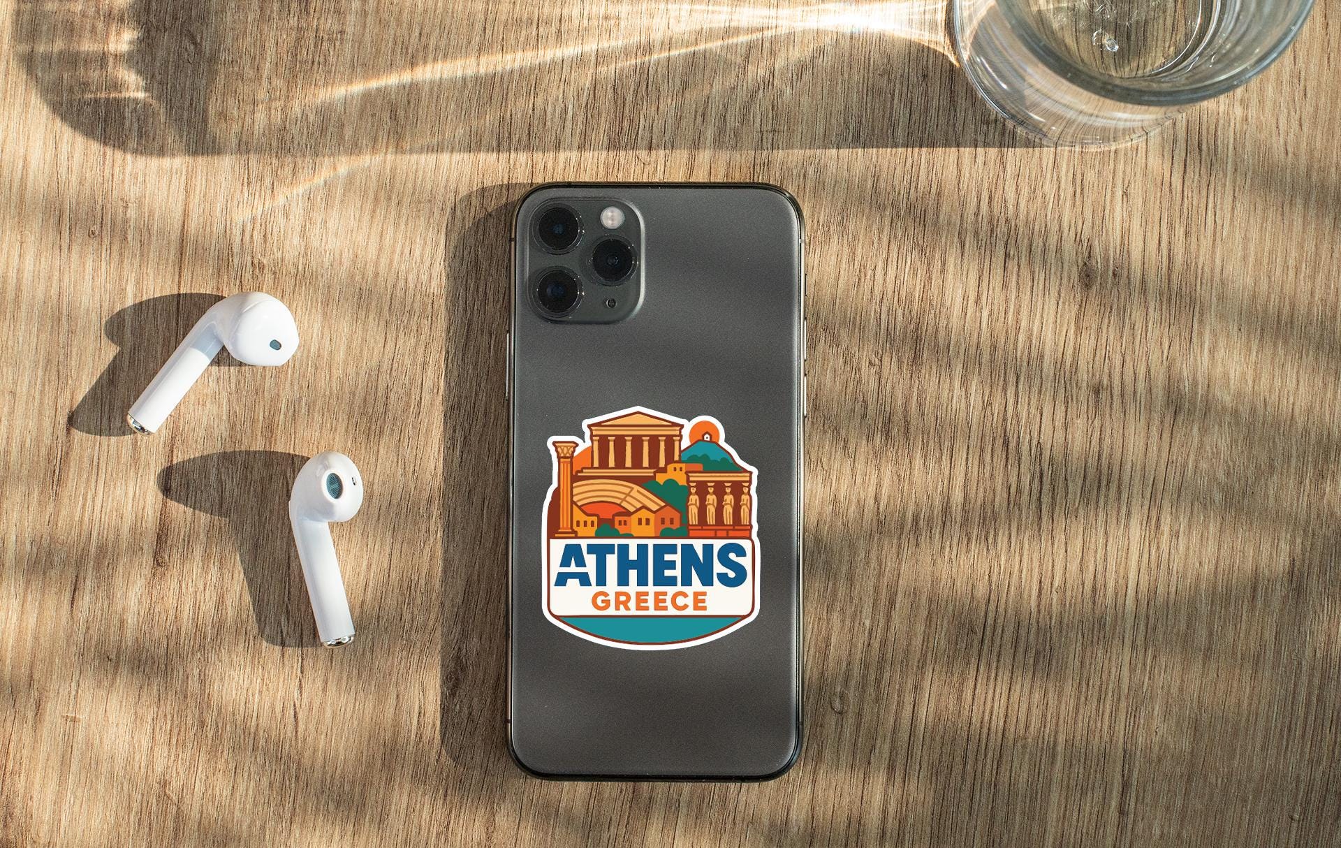 a smartphone with a sticker of Athens Greece on it, placed on a wooden surface alongside a pair of white earbuds and a glass of water.