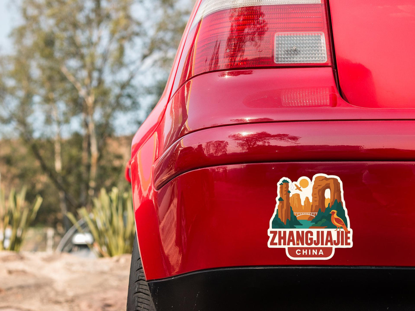 the rear end of a red car with a sticker on it, featuring a scenic landscape of mountains and a river.