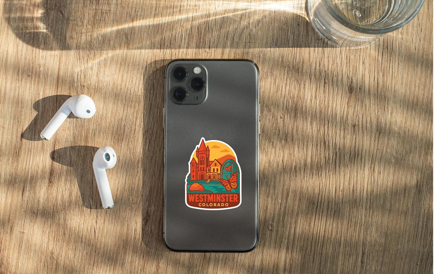 a smartphone with a sticker on the back, placed on a wooden surface alongside a pair of white earbuds and a glass of water.