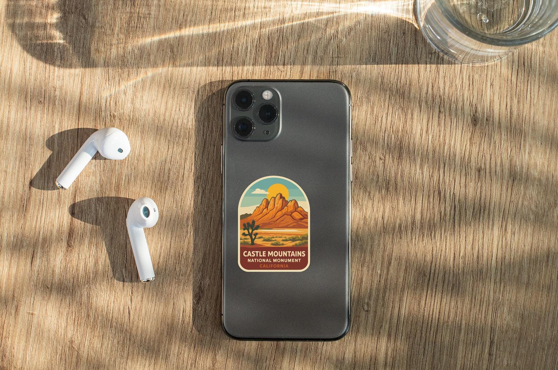 a smartphone with a sticker of a desert landscape on it, placed on a wooden surface alongside a pair of white earbuds and a glass of water.