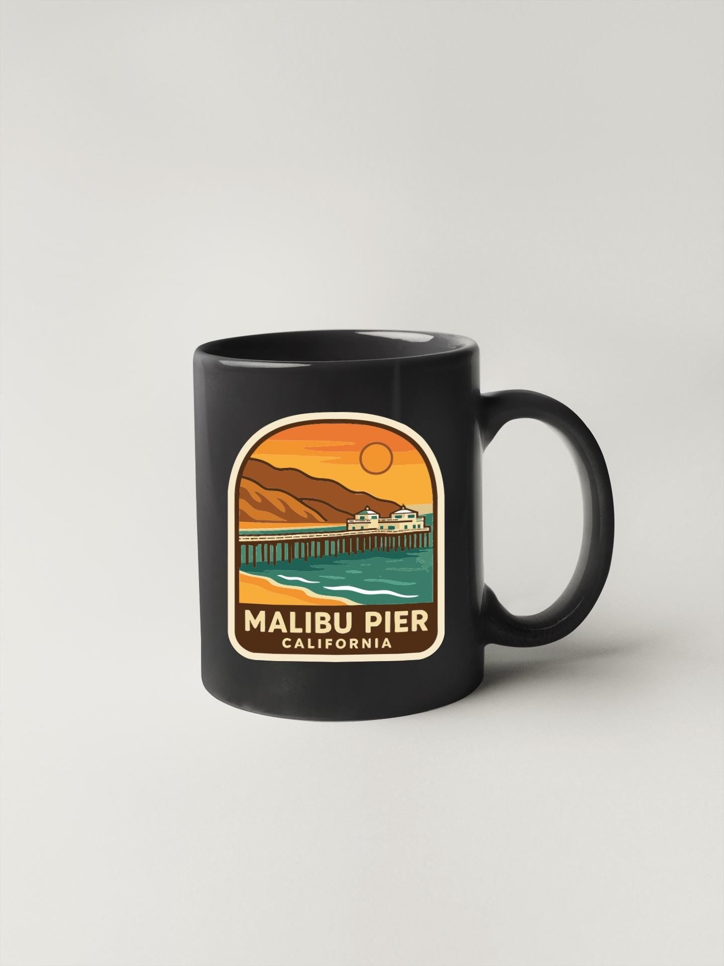 A black coffee mug with a graphic design depicting the Malibu Pier in California.