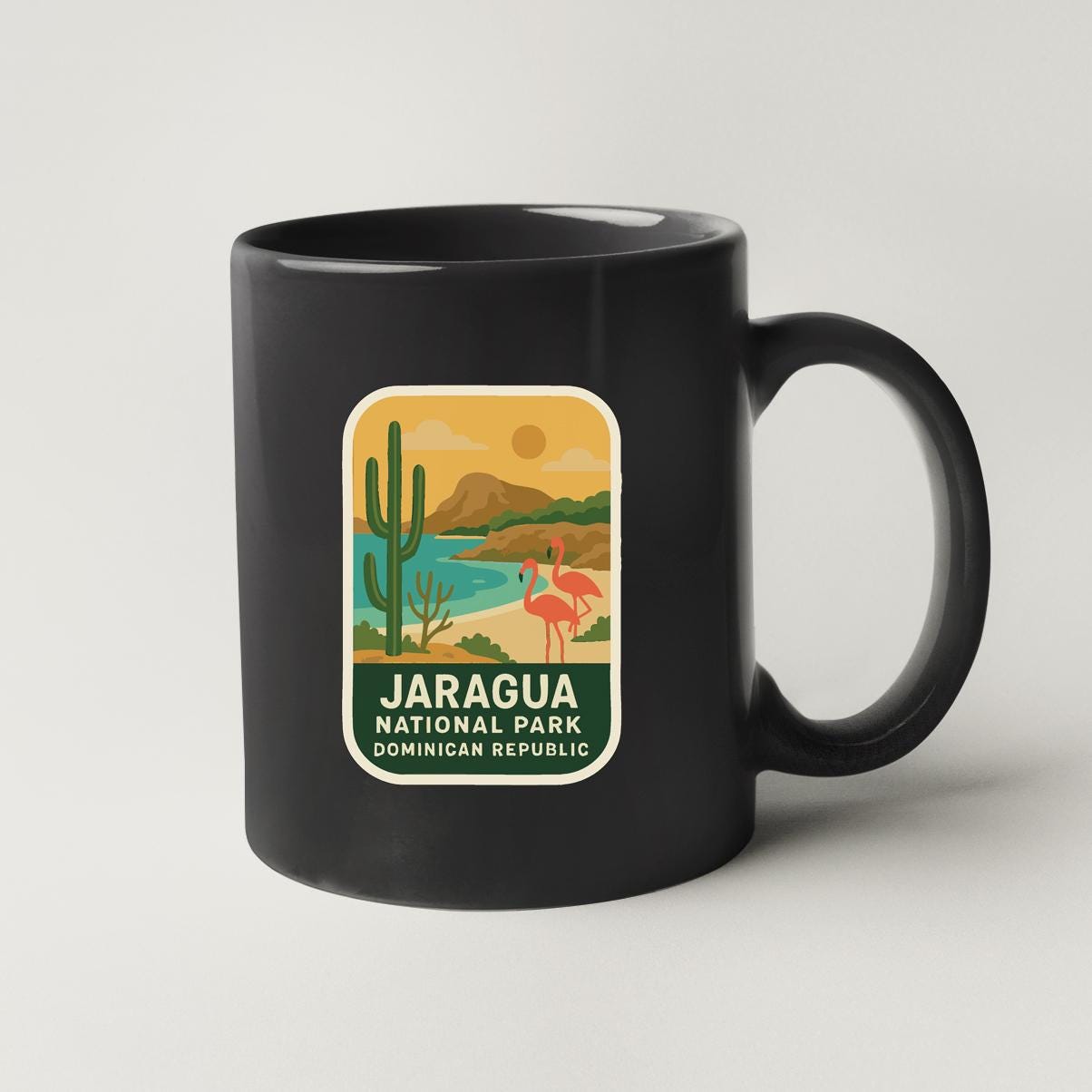 A black coffee mug with a colorful label that features a desert landscape with a cactus, flamingo, and mountains in the background, along with the text "JARAGUA NATIONAL PARK DOMINICAN REPUBLIC".