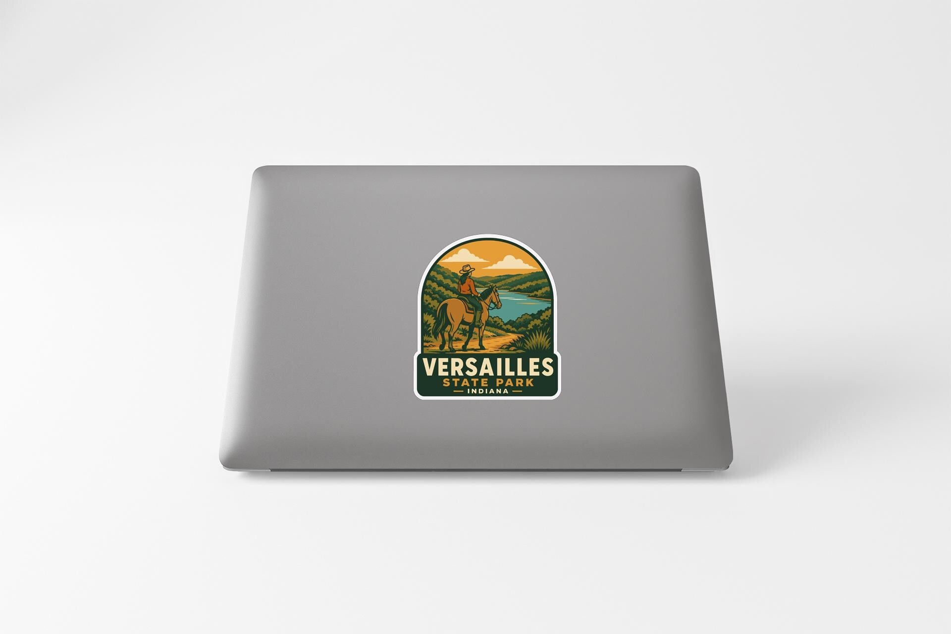 A sticker on the back of a laptop, depicting a scenic landscape with mountains, a river, and a person riding a horse.