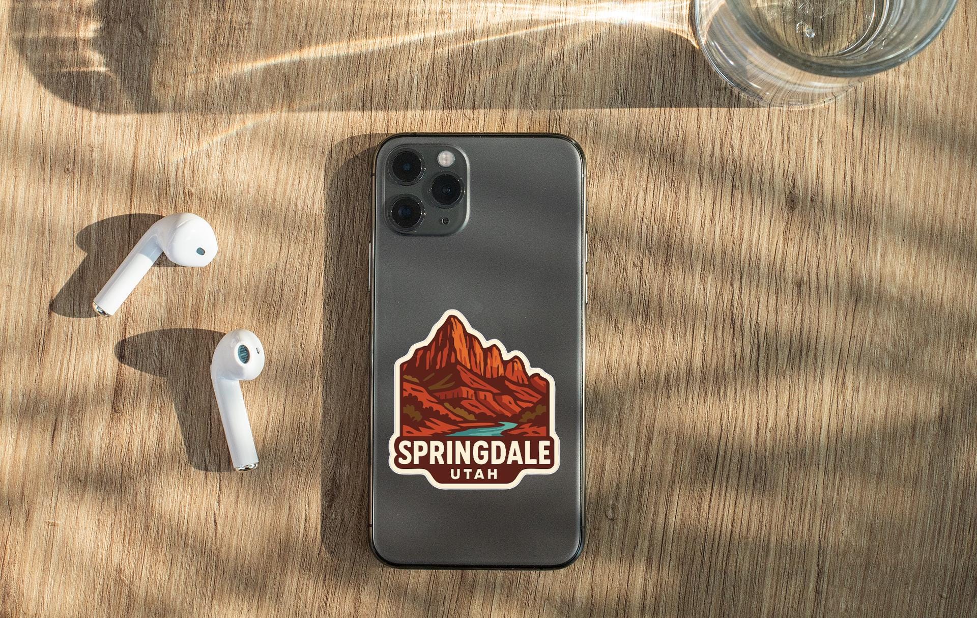 a smartphone with a sticker of a mountain range on it, along with a pair of white earbuds and a glass of water on a wooden surface.