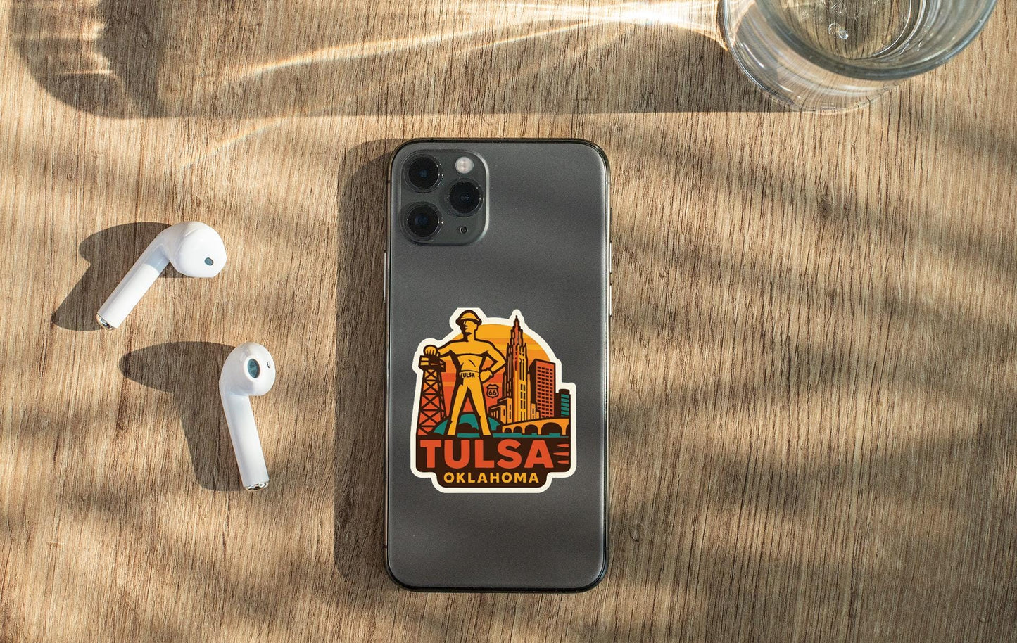 a smartphone with a sticker of a city skyline and the text "Tulsa Oklahoma" on its back, along with a pair of white earbuds and a glass of water on a wooden surface.