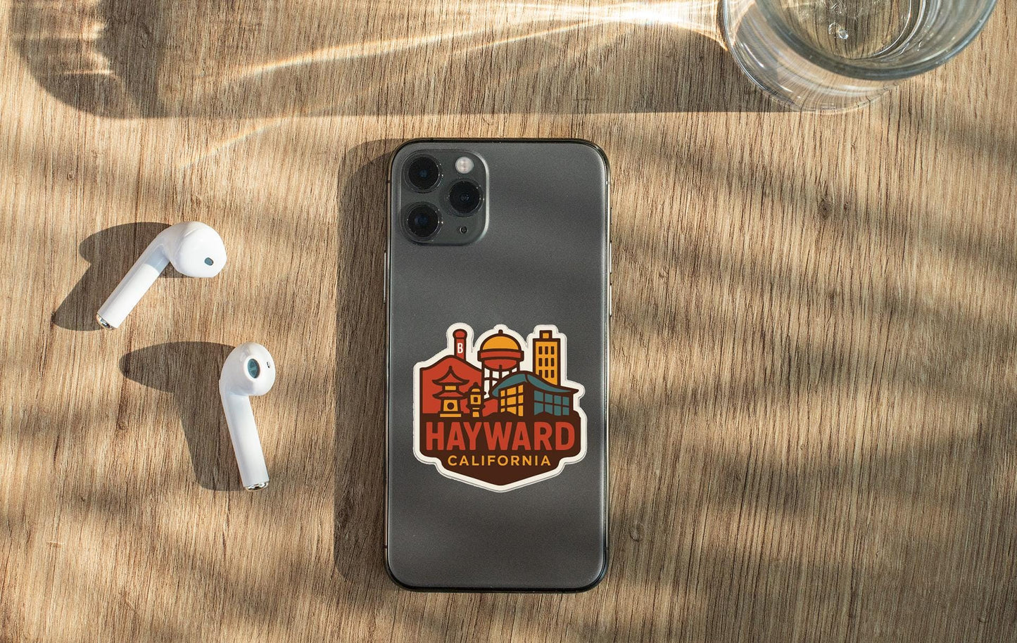 a smartphone with a sticker of the town of Hayward, California, placed on a wooden surface alongside a pair of white earbuds and a glass of water.