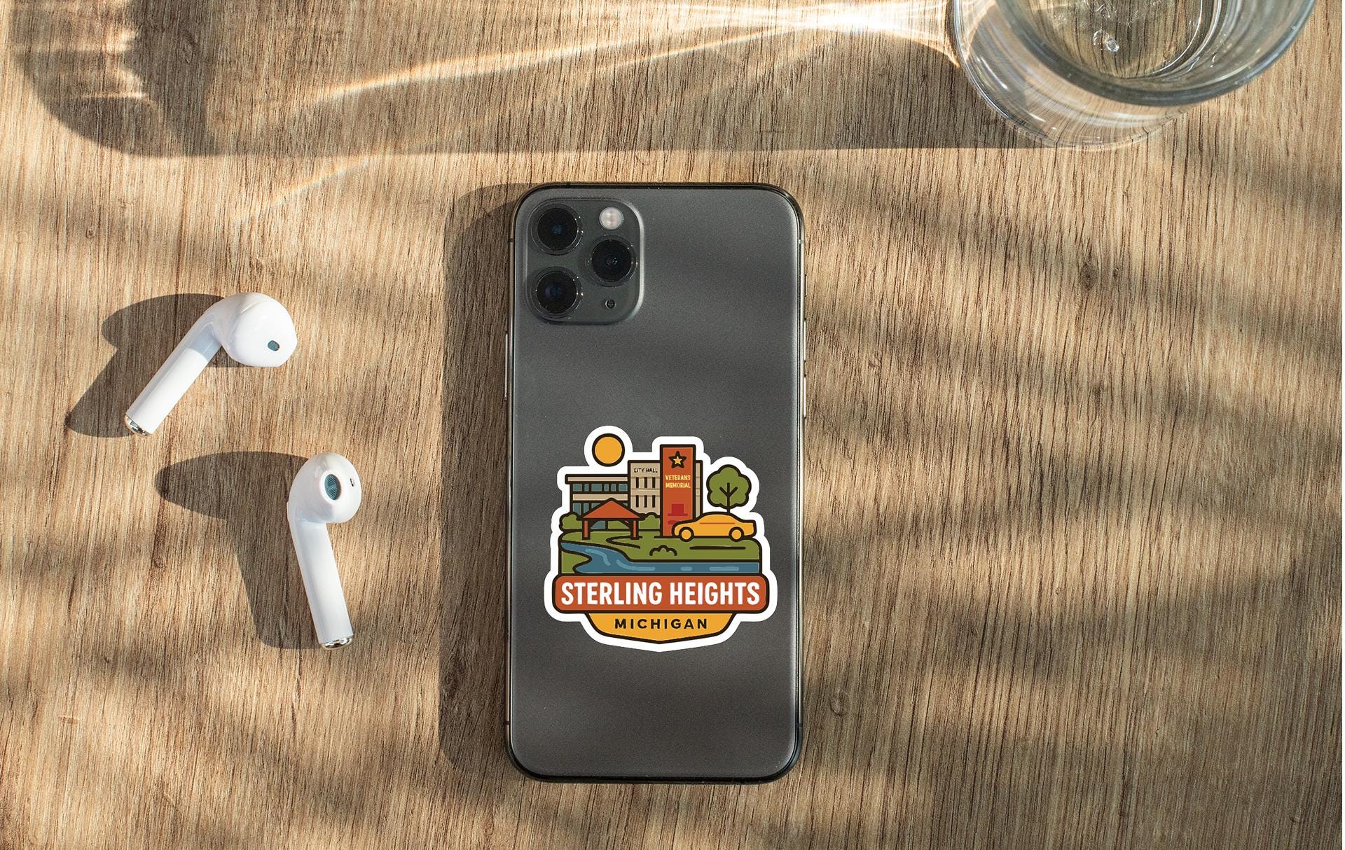 a smartphone with a sticker on the back, placed on a wooden surface alongside a pair of white earbuds and a glass of water.