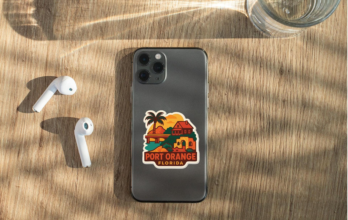 a smartphone with a sticker of a tropical beach and palm trees, along with a pair of white earbuds and a glass of water on a wooden surface.