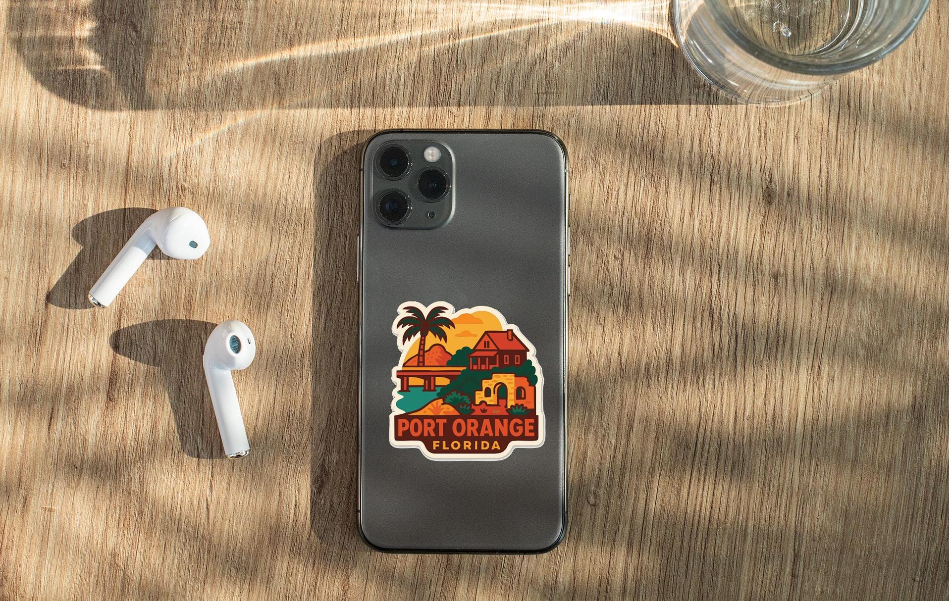 a smartphone with a sticker of a tropical beach and palm trees, along with a pair of white earbuds and a glass of water on a wooden surface.