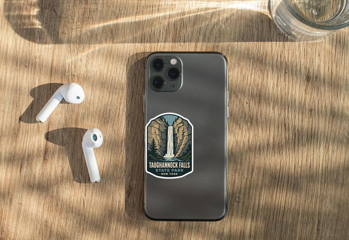 a smartphone with a sticker of Taughannock Falls on its back, along with a pair of white earbuds and a glass of water on a wooden surface.
