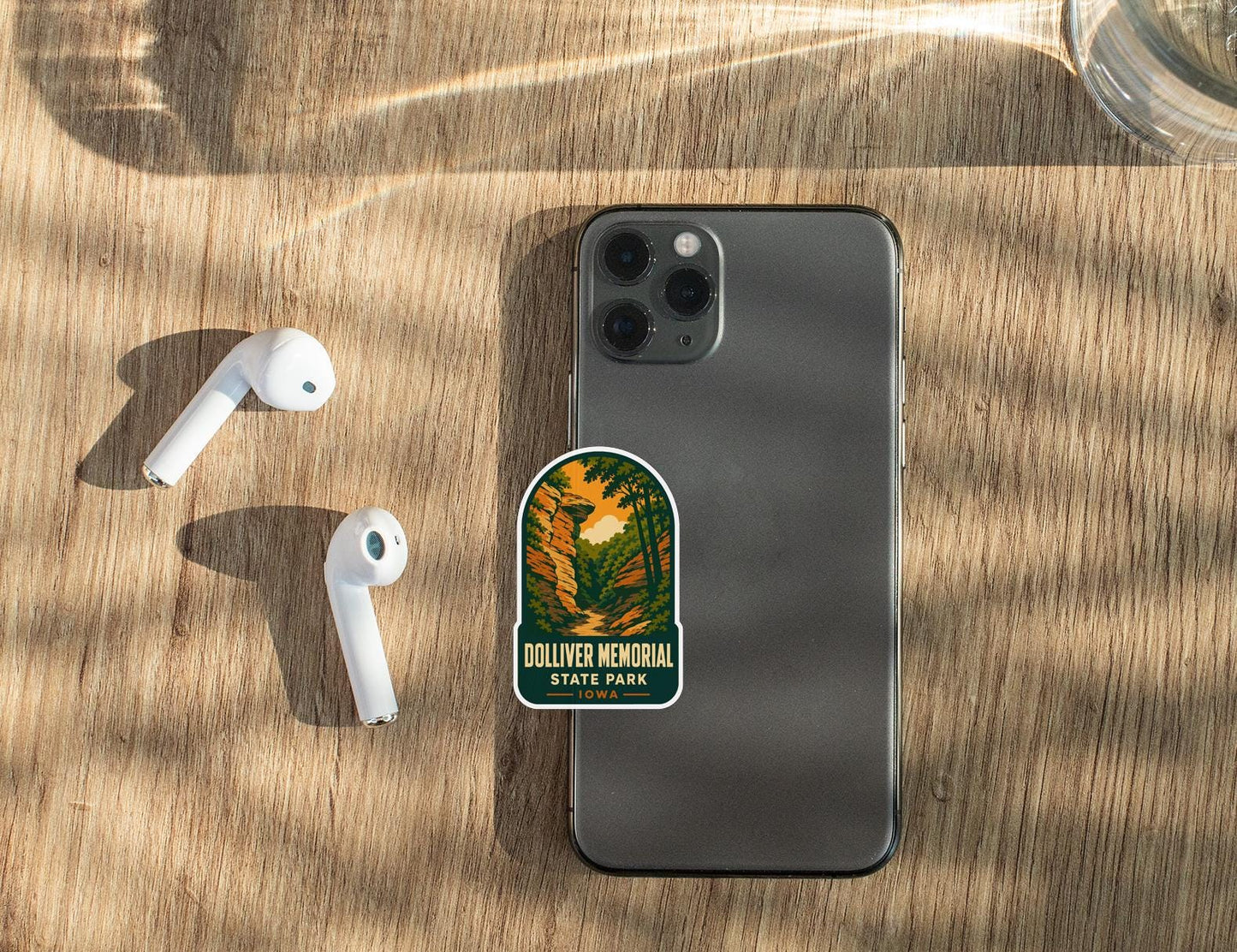 a smartphone with a sticker on it, along with a pair of white earbuds and a glass of water on a wooden surface.