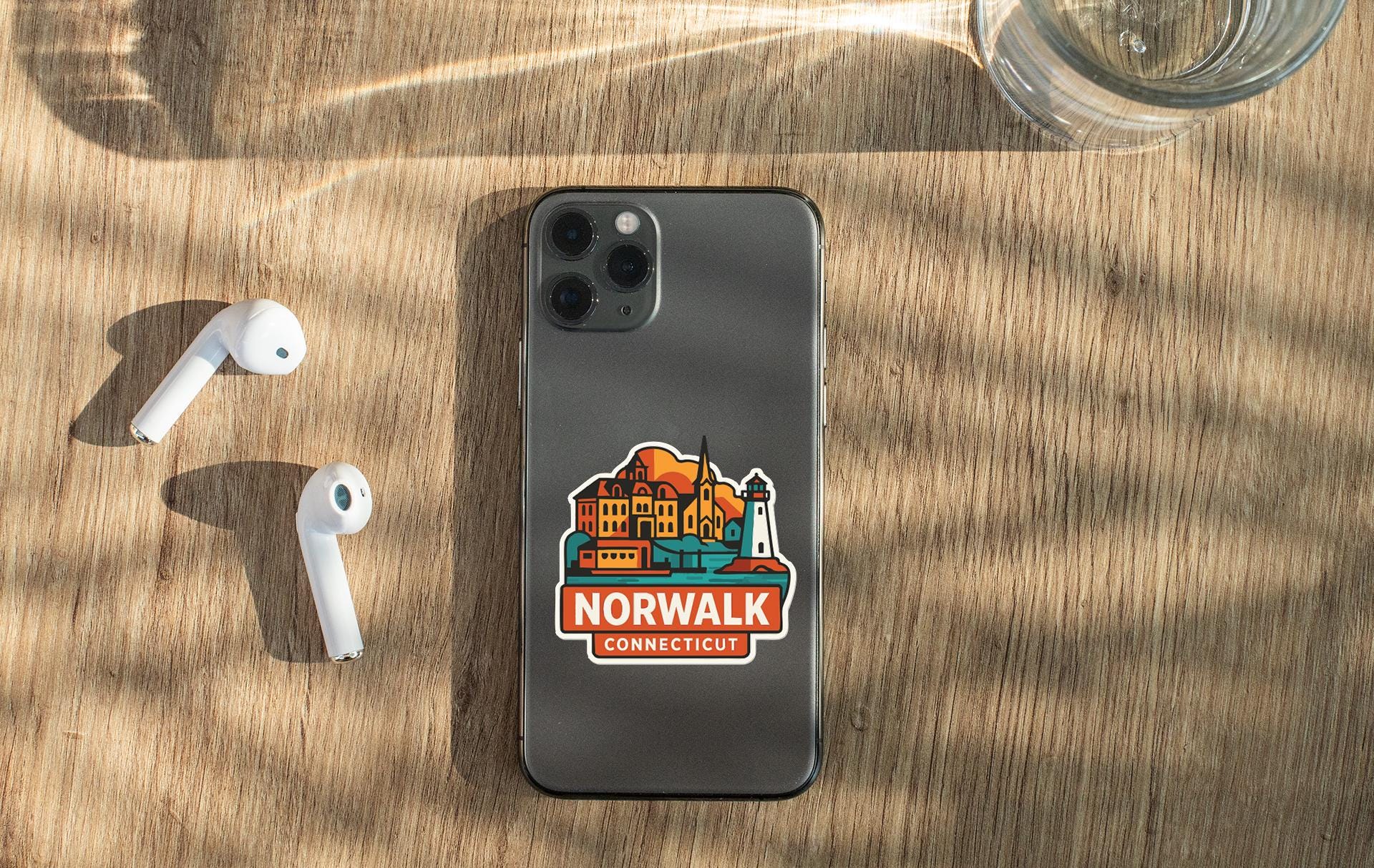 a smartphone with a sticker on it, placed on a wooden surface alongside a pair of white earbuds and a glass of water.
