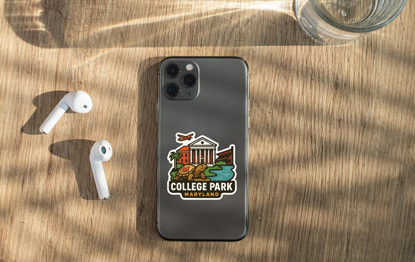 a smartphone with a sticker on it, placed on a wooden surface alongside a pair of white earbuds and a glass of water.