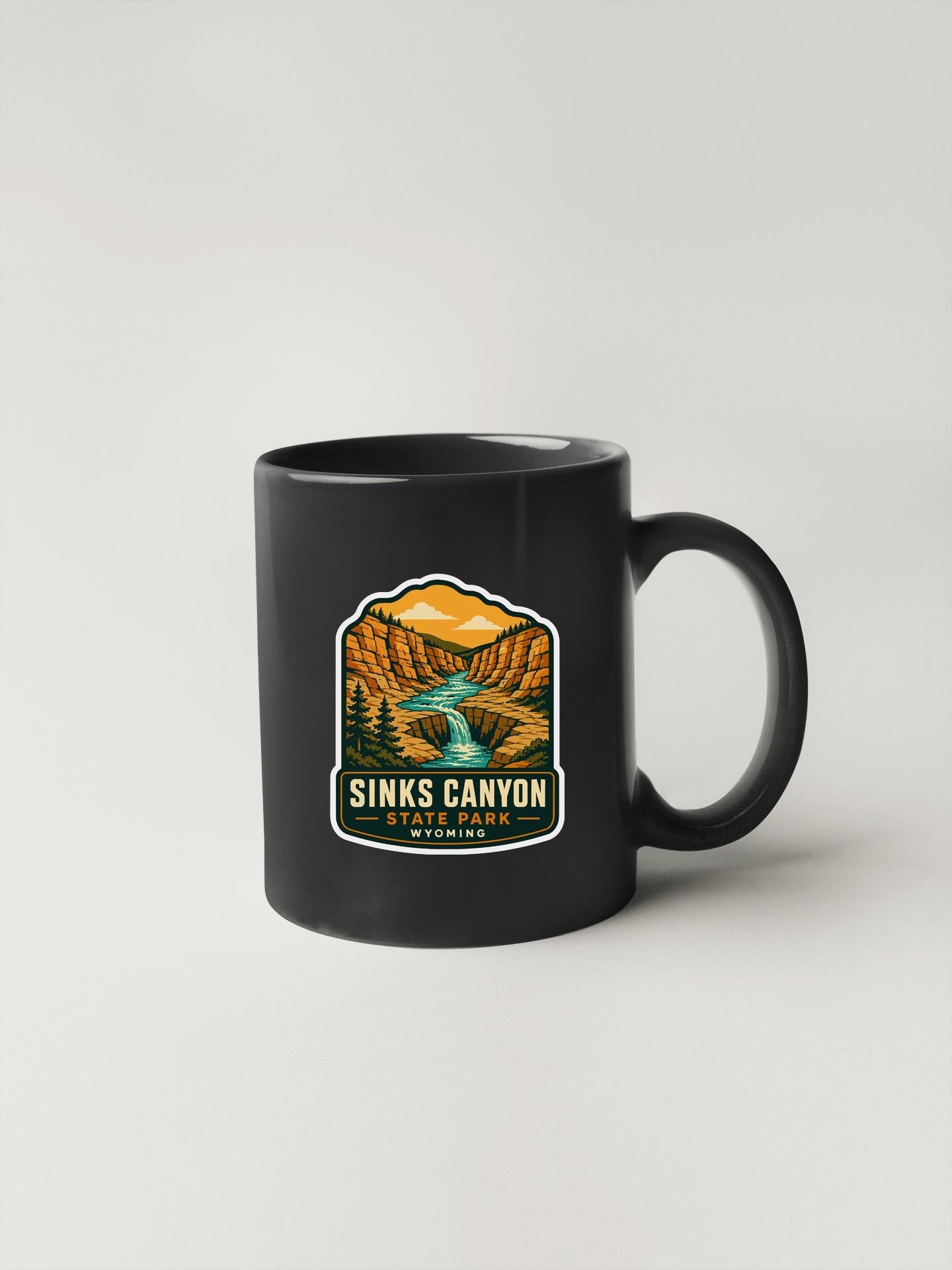 A black coffee mug with a graphic design depicting a scenic landscape, including a waterfall and mountains, along with the text "SINKS CANYON" and "YOHAWA PARK, WYOMING".